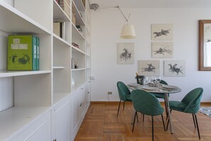 Un comedor con una mesa redonda, sillas verdes y una estantería blanca llena de libros.