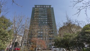 Um edifício residencial alto com várias varandas, rodeado por árvores e carros estacionados.
