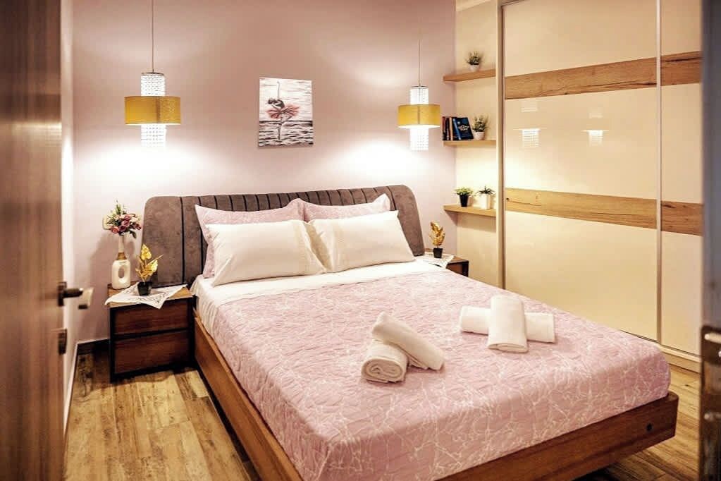 A neatly made bed with a pink bedspread, two bedside lamps, a nightstand with a vase of flowers, and a wooden wardrobe with a mirror.