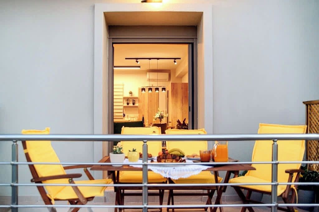A balcony with yellow chairs and a table set for dining, overlooking a modern kitchen with wooden cabinets and a dining area.