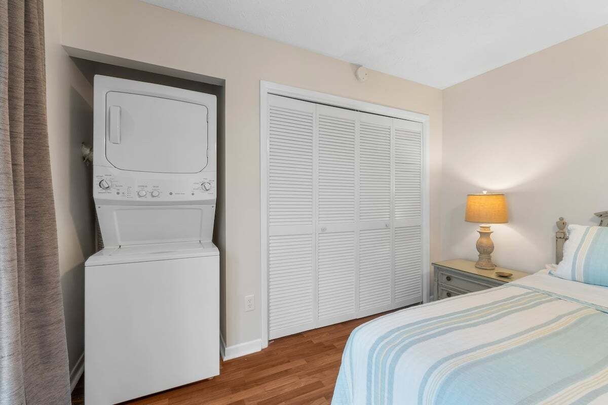 A bedroom with a bed, a nightstand with a lamp, a washing machine, and a set of white louvered closets.