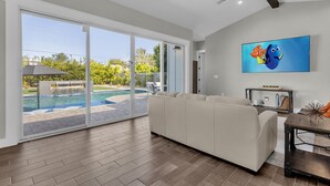 Un salón amplio con un gran ventanal corredera que da a una zona de piscina, un sofá blanco y un televisor de pantalla plana en la pared.