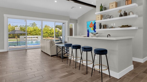 Una cocina moderna con armarios blancos, una zona de bar con taburetes azules y vistas a una piscina.