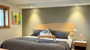 Une chambre avec un grand lit, une table de chevet ornée d’un vase et une fenêtre avec des stores.