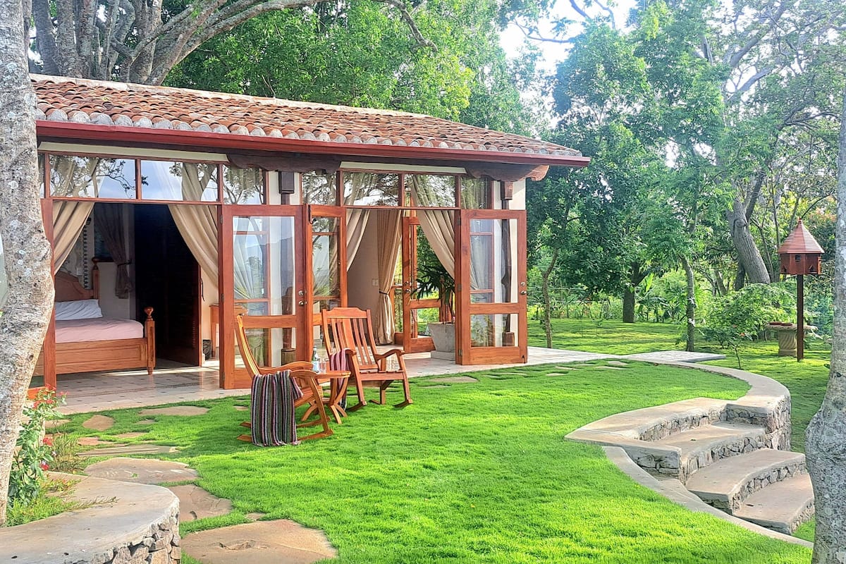 A cozy, single-story wooden structure with large windows and a balcony, surrounded by lush greenery and a stone pathway.