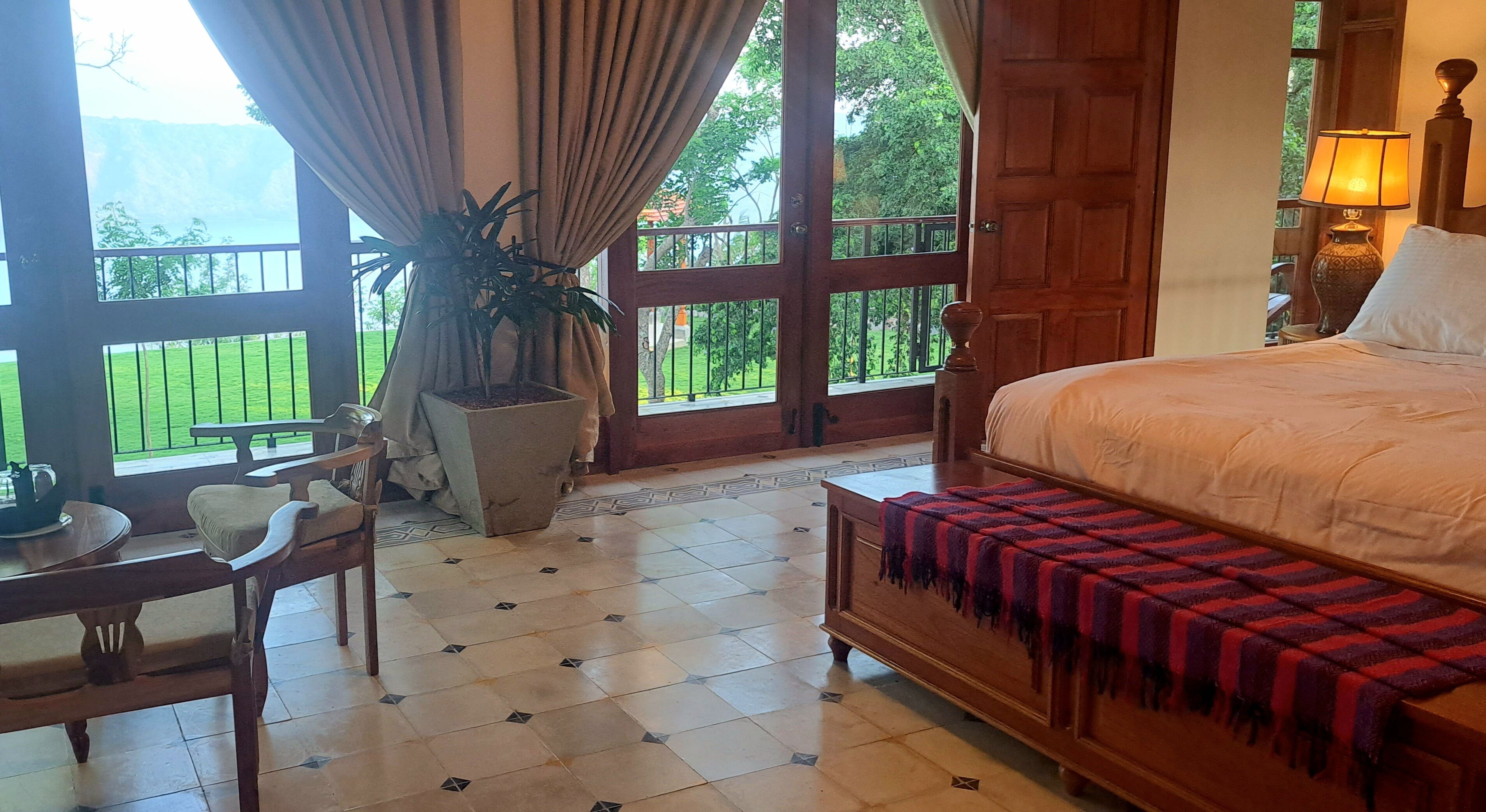 A hotel room with a bed, a table with chairs, a potted plant, and large windows with a view of greenery.