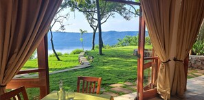 A view from a room with a table and chairs, overlooking a lush green landscape with a pathway leading to a body of water and hills in the distance. - El Guayacán (Masaya)