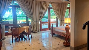 Una habitación con cama, una mesa con sillas, una lámpara y vistas al exterior a través de amplios ventanales.