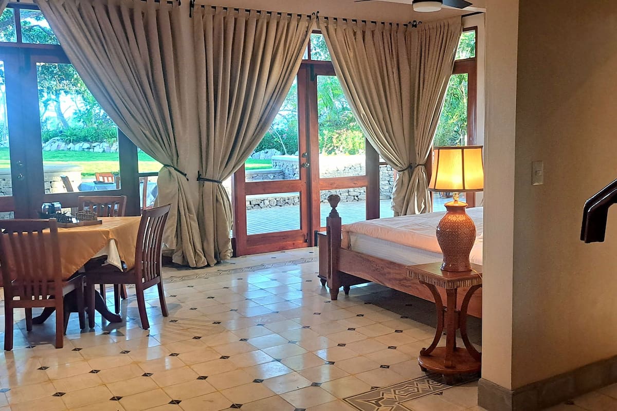 A room with a bed, a table with chairs, a lamp, and a view of the outdoors through large windows.