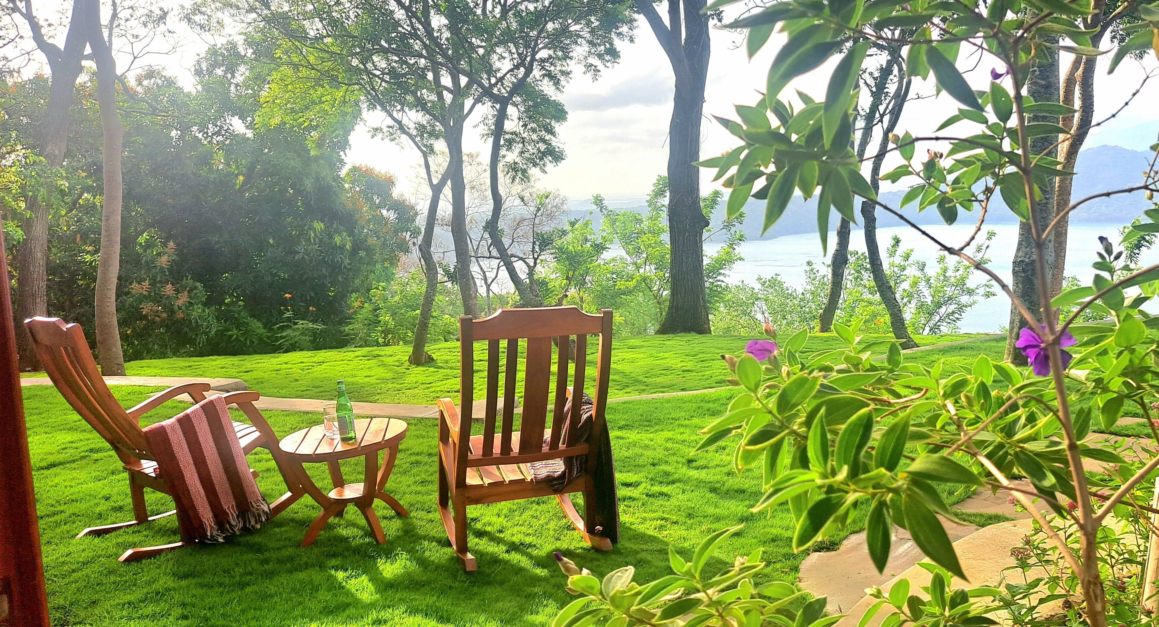 A grassy outdoor area with two wooden chairs and a table, surrounded by trees and a view of a body of water.