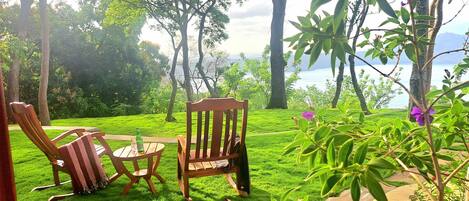 A grassy outdoor area with two wooden chairs and a table, surrounded by trees and a view of a body of water.