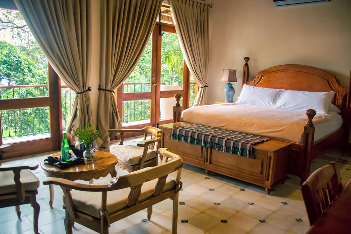 A bedroom with a wooden bed, a small table with a plant, and a view of greenery through the window.