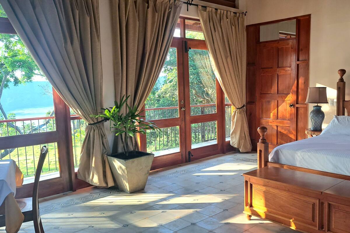 A bedroom with a wooden bed, a potted plant, a table, and a view of the outdoors.