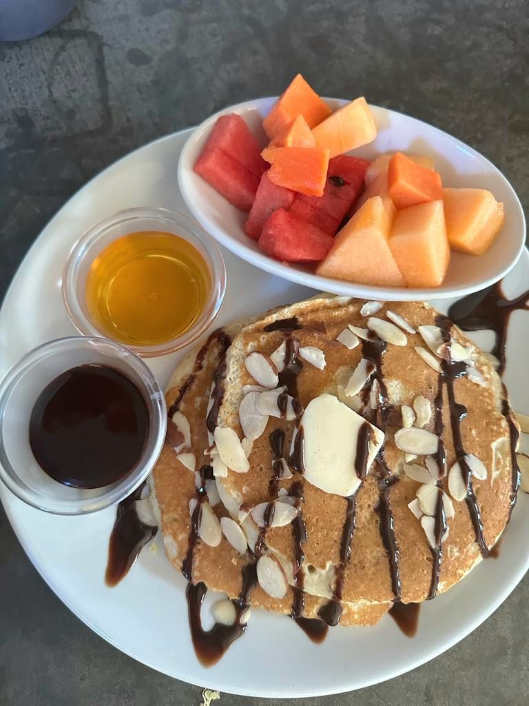 A plate with pancakes topped with almonds and chocolate syrup, accompanied by two small bowls of syrup and a bowl of cubed melon.