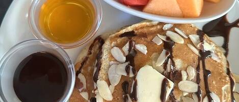 A plate with pancakes topped with almonds and chocolate syrup, accompanied by two small bowls of syrup and a bowl of cubed melon.