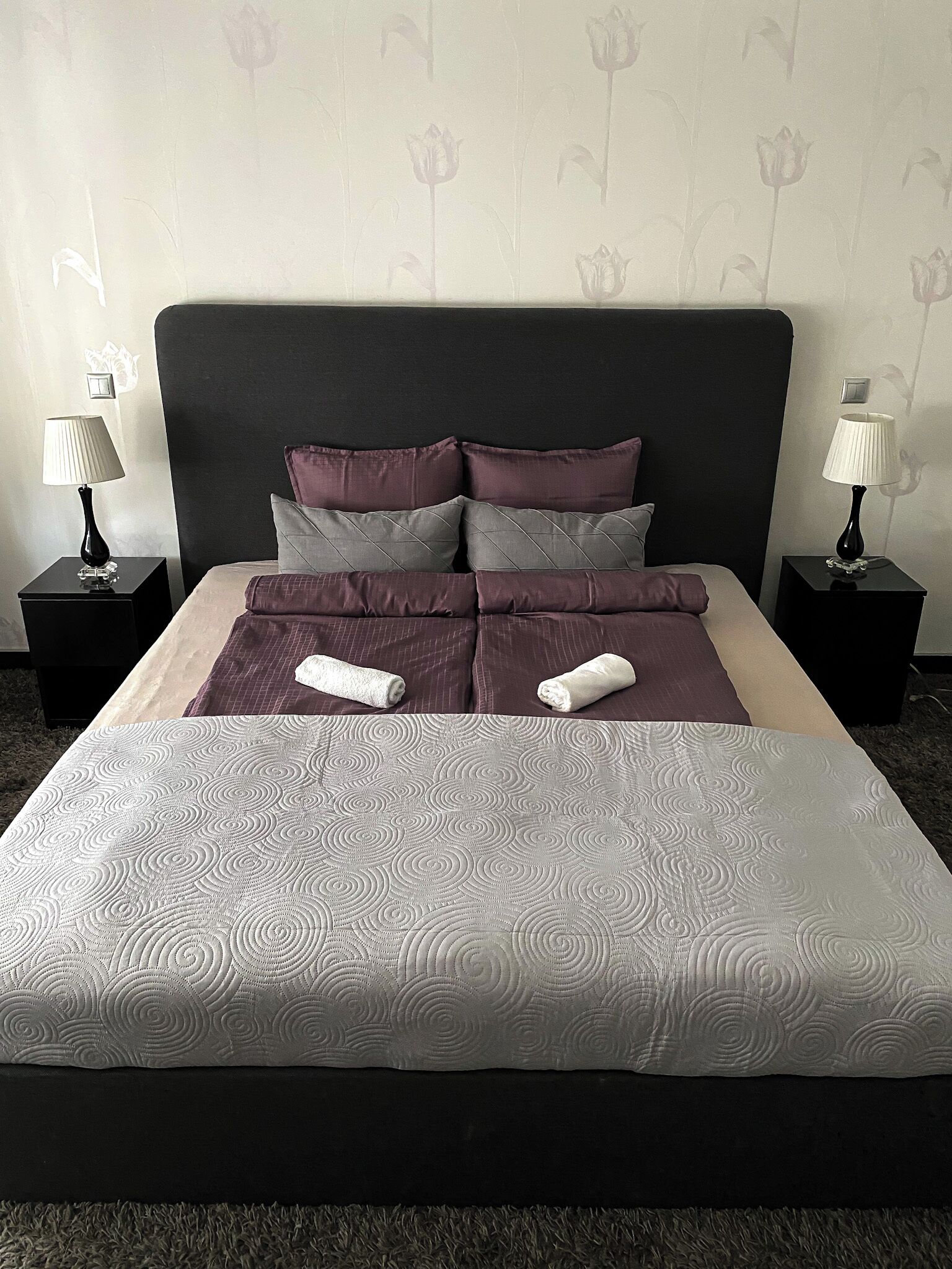 Un lit bien fait, recouvert d’une couvre-lit à motifs, avec deux oreillers et deux serviettes enroulées. De chaque côté du lit, on trouve une table de chevet sur laquelle est posée une lampe.