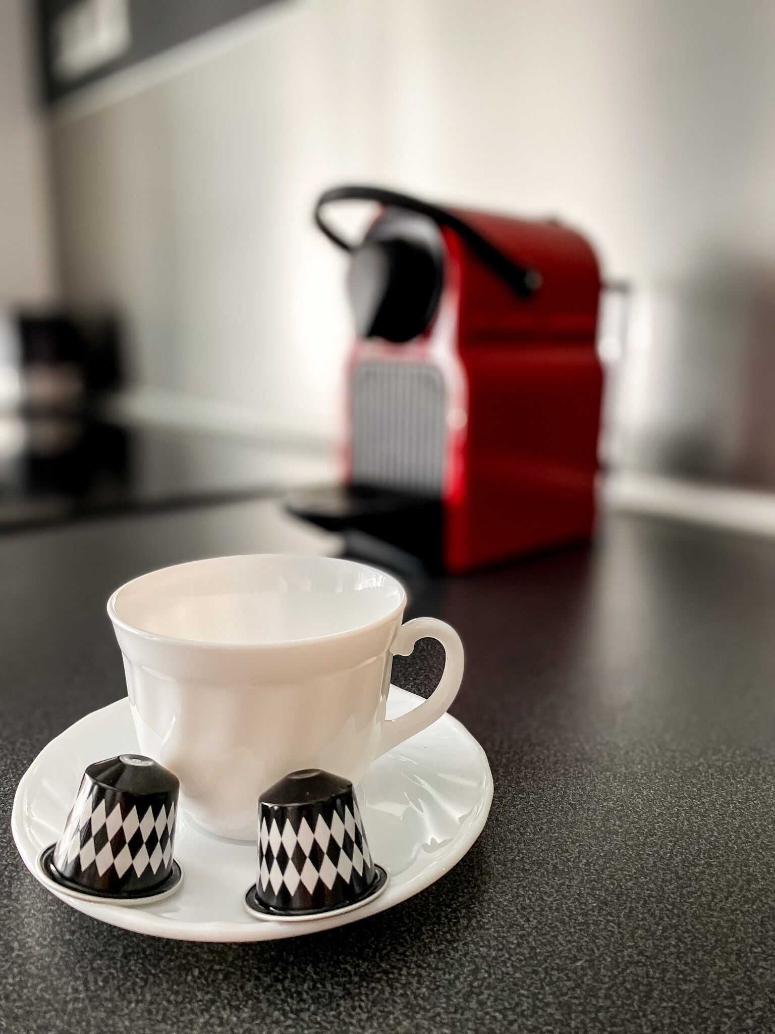 A white cup with a saucer and two black and white patterned sugar cubes on a dark countertop. A red and black object is in the background.