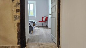 A hallway with a bed, a red chair, and a door leading to another room. - Maison de Campagne au Coeur du Parc Naturel Regional des Monts D'ardeche (Issamoulenc)