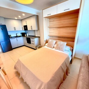 A bedroom with a bed, a wooden headboard, a kitchen with white cabinets, and a refrigerator.