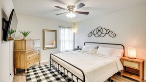 A bedroom with a bed, a wooden dresser, a ceiling fan, a nightstand with a lamp, a mirror, and a window with curtains. - Palm Coast Duplex w/ Screened Patio, Near Beaches! (Palm Coast)