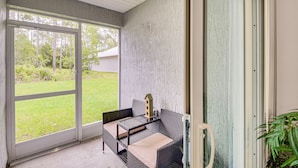 A room with a glass door leading to a grassy yard, a wicker sofa with a glass table, and a small decorative item on the table. - Palm Coast Duplex w/ Screened Patio, Near Beaches! (Palm Coast)