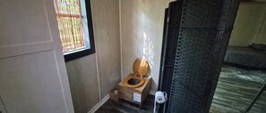 A small bathroom with a wooden toilet, a window with a metal screen, and a shower area with a black mat.