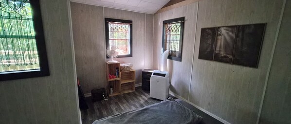 A room with a bed, a small desk, a chair, a radiator, and a window with a view of greenery.