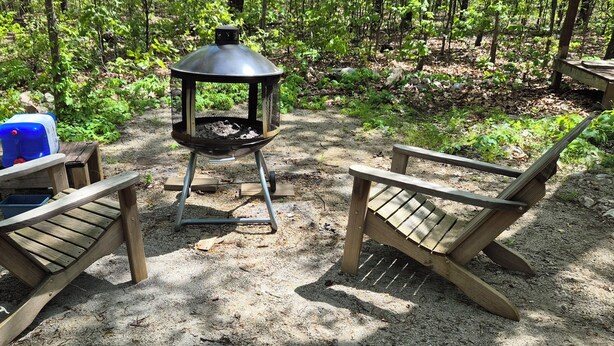 Un camping con dos sillas de madera, una parrilla portátil y un recipiente azul.