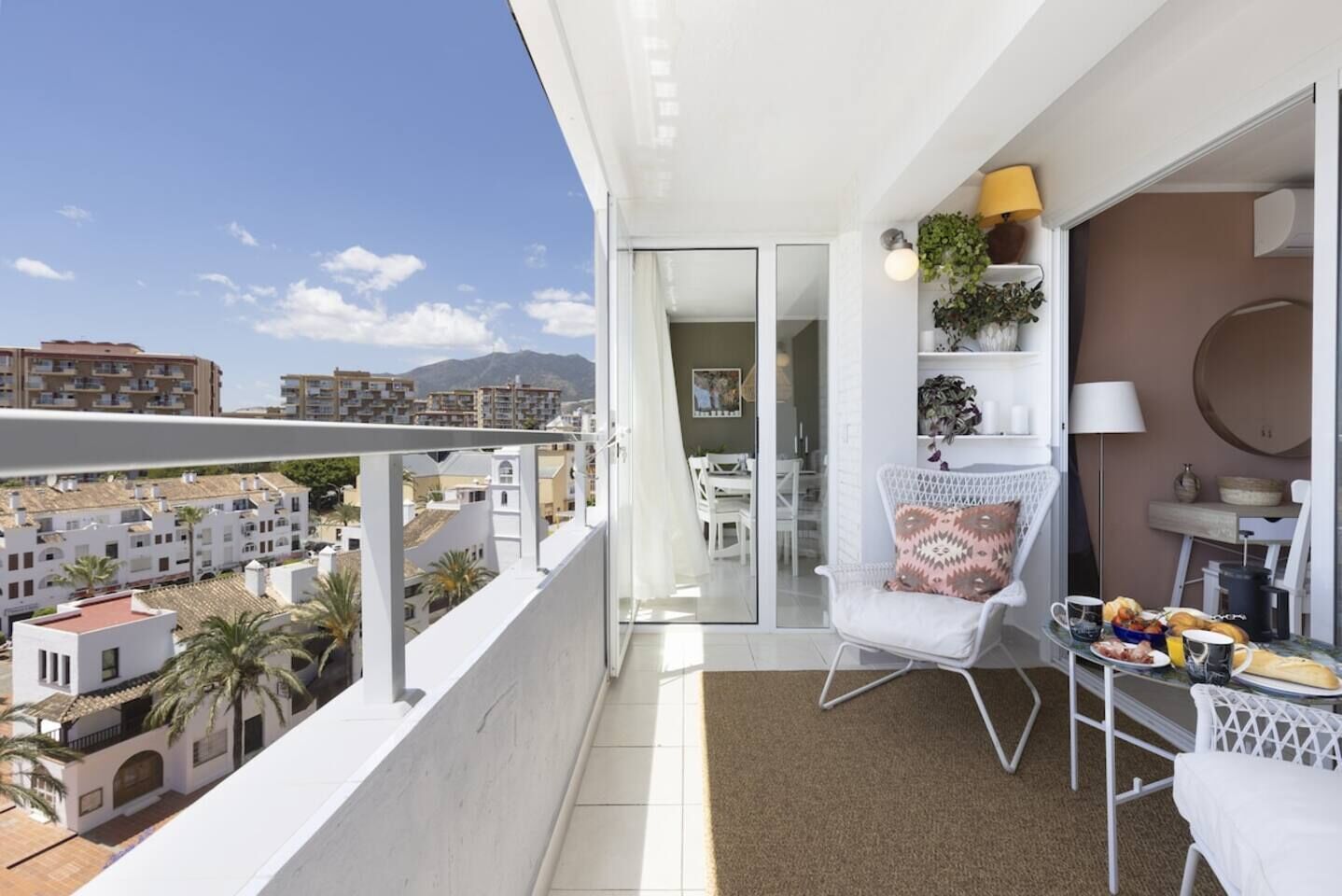 Un balcón con una silla blanca, una mesita con cosas para el desayuno y vistas a la ciudad.