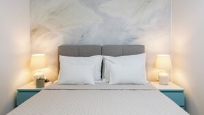 Ein Bett mit weißer Bettwäsche, zwei weiße Kissen und ein grauer Kopfteil. Auf den beiden Nachttischen stehen Lampen und jeweils eine kleine Pflanze.