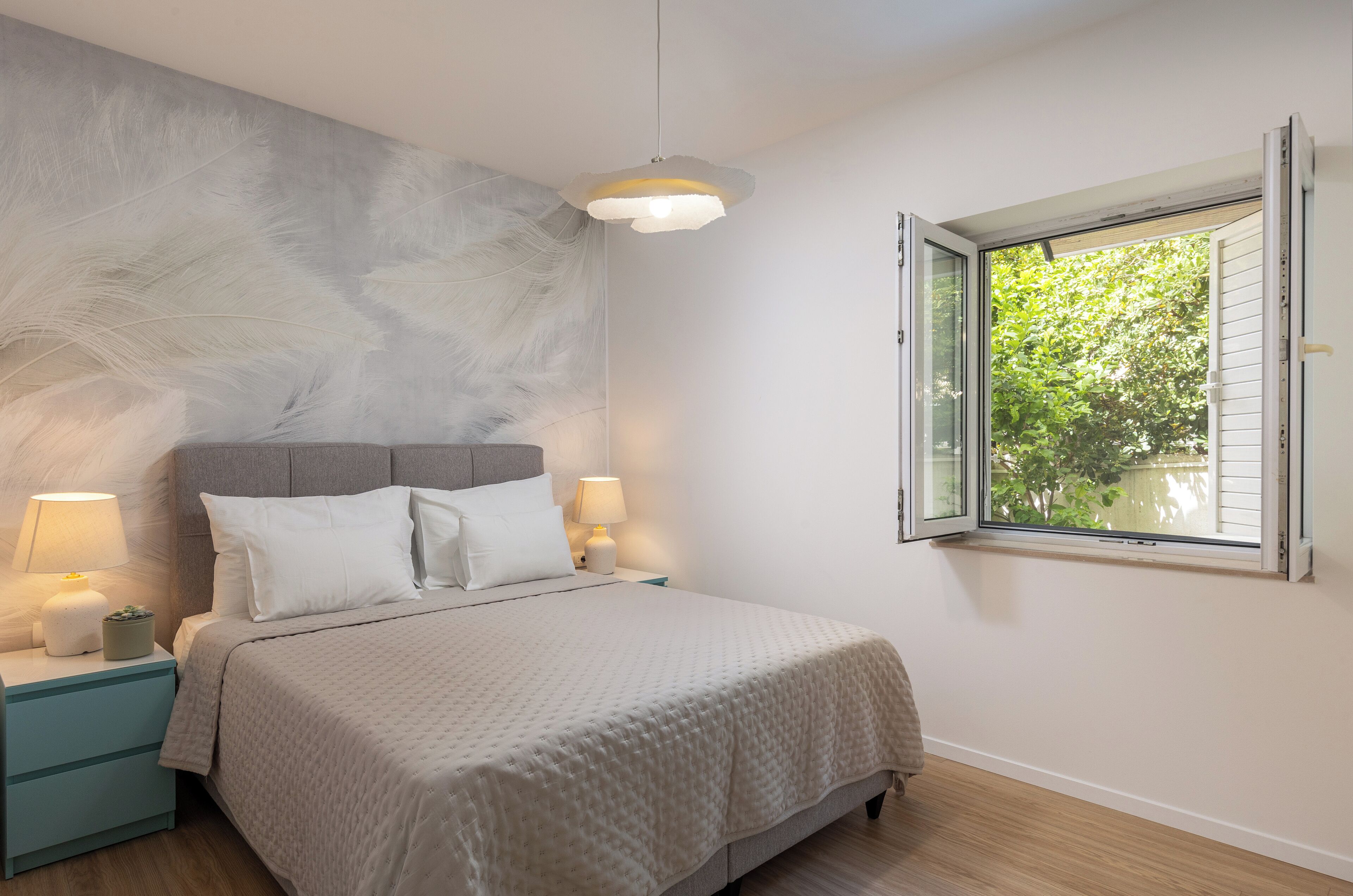 A bedroom with a bed, two bedside lamps, a nightstand, a window with a view of greenery, and a textured wall.