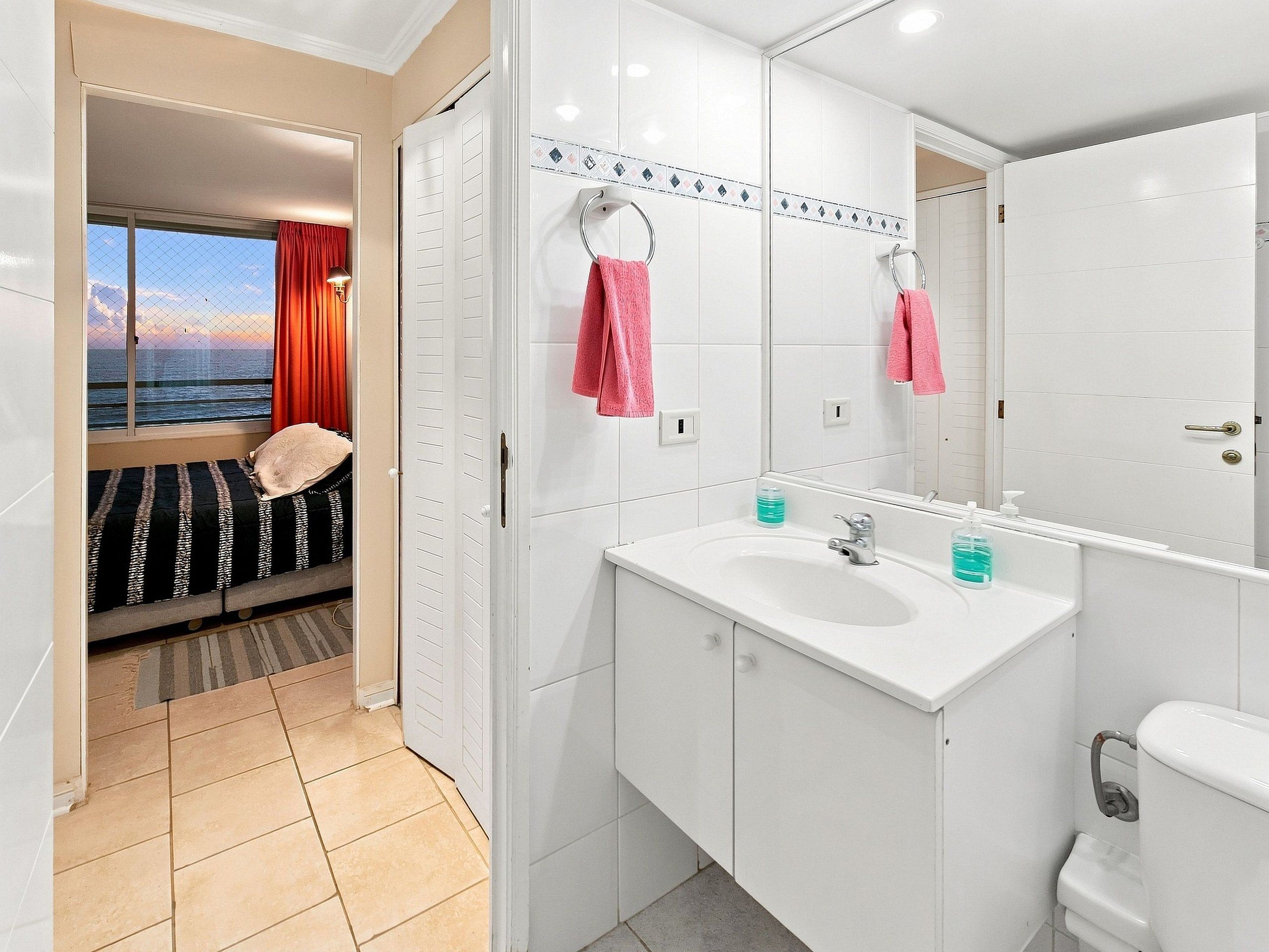 Een badkamer met een wit wastafel, spiegel en toilet. Er is een raam met uitzicht op zee.