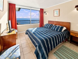 Habitación de hotel con cama, escritorio, silla, ventana con vista al mar y un cuadro en la pared.