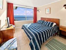 Habitación de hotel con cama, escritorio, silla, ventana con vista al mar y un cuadro en la pared.