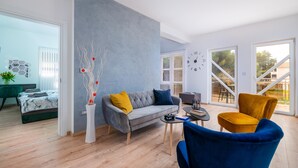 Un salon moderne avec un canapé gris, des coussins jaunes et bleus, une table basse et un fauteuil bleu. Une grande fenêtre offre une vue sur les arbres et donne sur un balcon.