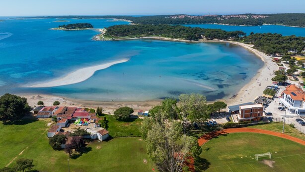 Vue aérienne d’une zone côtière avec une plage, des eaux d’un bleu limpide et un petit village composé de maisons et d’un terrain de football.