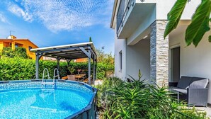 Ein Schwimmbecken mit überdachtem Bereich, umgeben von Grünflächen und einem modernen Gebäude mit Steinverzierungen.