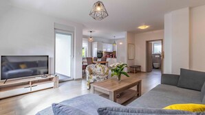 Ein modernes Wohnzimmer mit Flachbildfernseher, einer Couch, einem Couchtisch und einem Essbereich im Hintergrund.