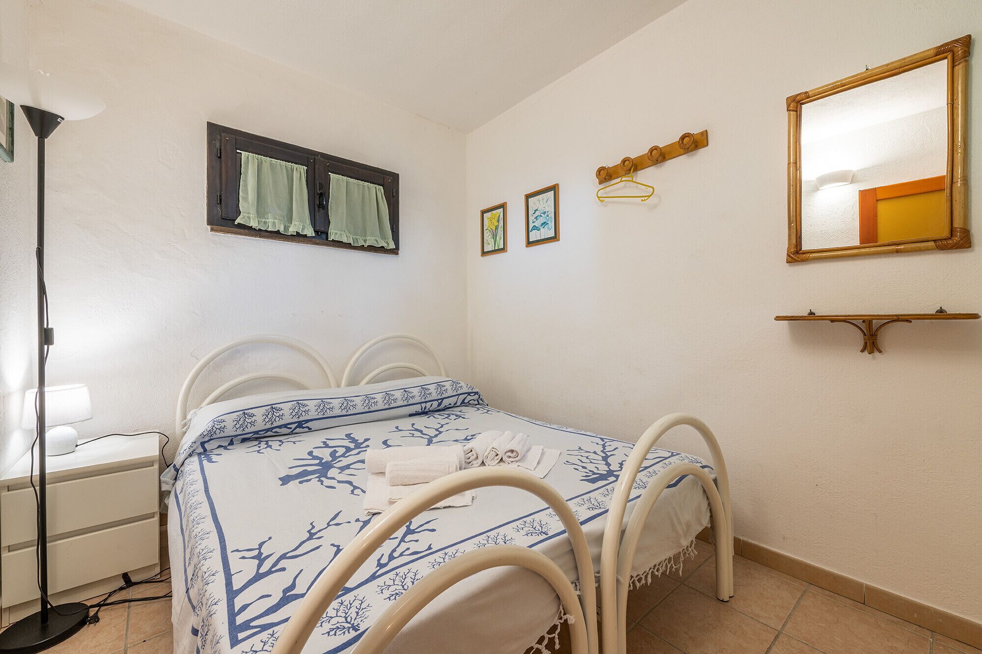 Un dormitorio con cama, mesita de noche, lámpara, ventana con cortinas, espejo en la pared y cuadros enmarcados.