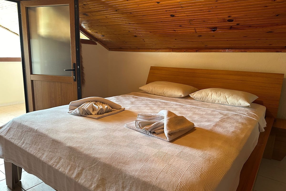Ein ordentlich bezogenes Bett mit Holz-Kopfteil, einem gefalteten Handtuch und einem Kissen auf einer strukturierten, beige farbenen Bettdecke.