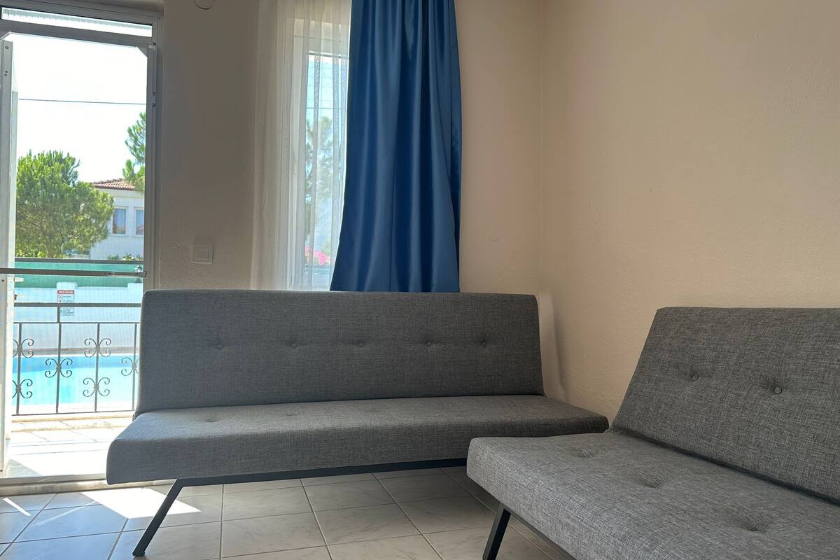Ein Zimmer mit zwei grauen Sofas, einem Fenster mit blauen Vorhängen und Blick auf einen Pool.