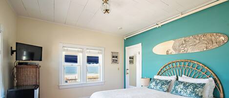 A bedroom with a bed, a television, a window with a view, and a surfboard on the wall.