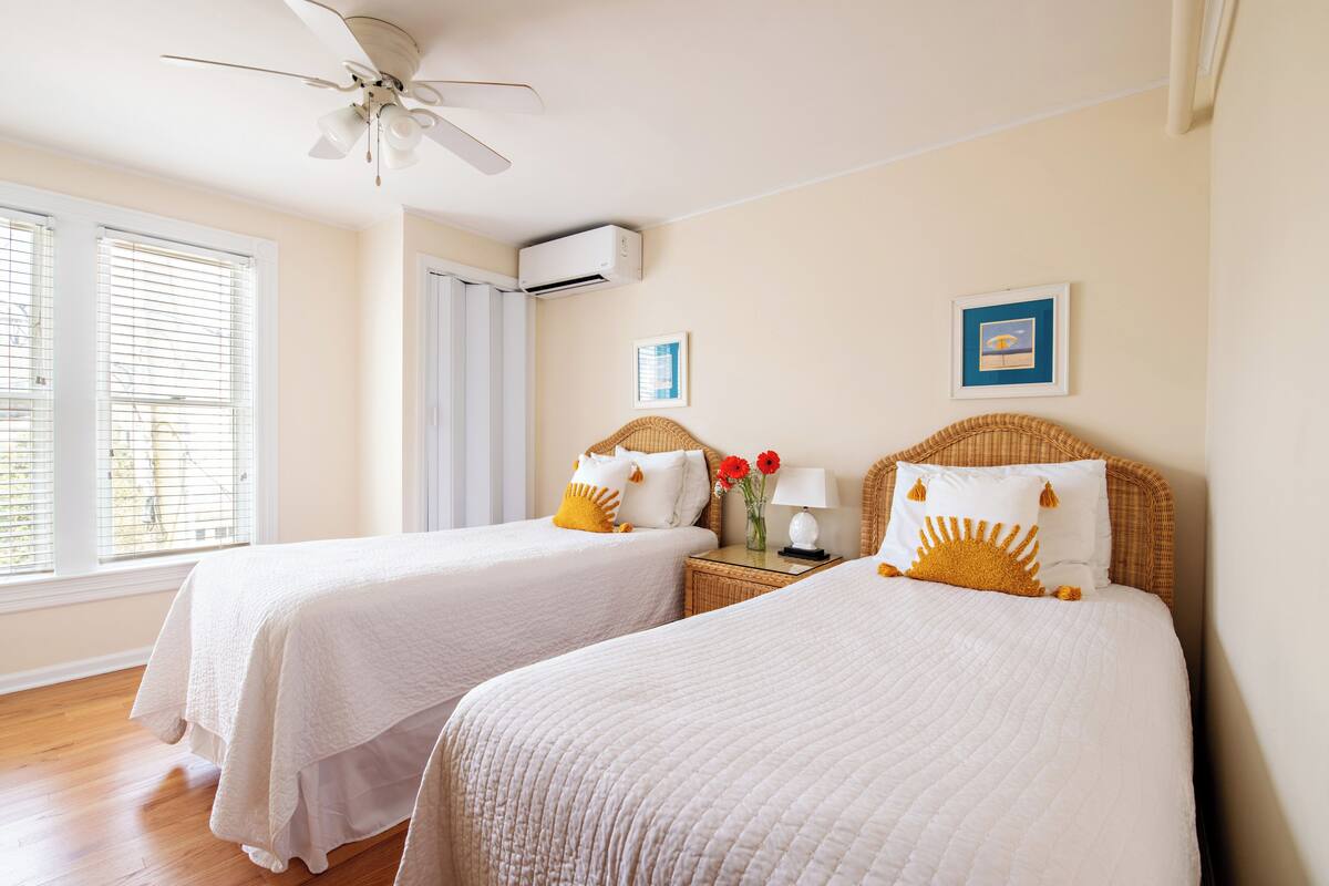 A hotel room with two beds, a ceiling fan, an air conditioning unit, and a window with blinds.