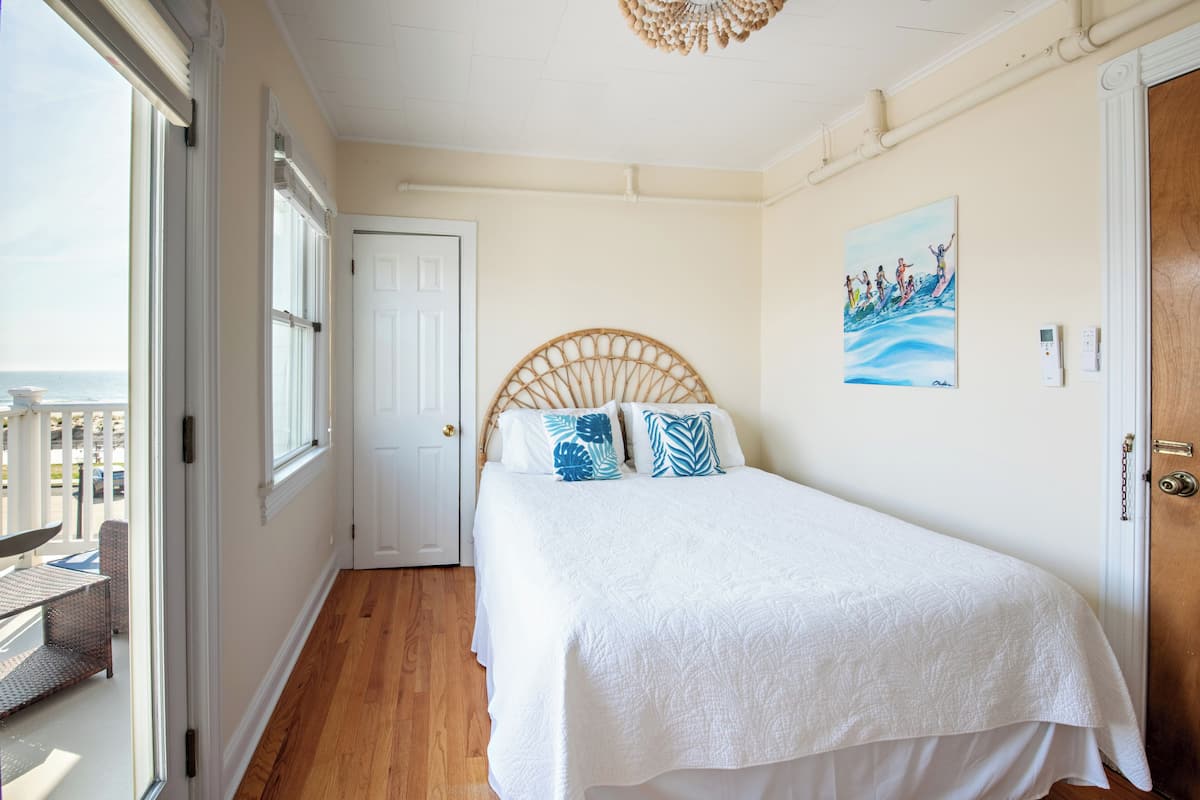 A bedroom with a bed, a wooden headboard, a white door, a painting on the wall, and a view through a window.