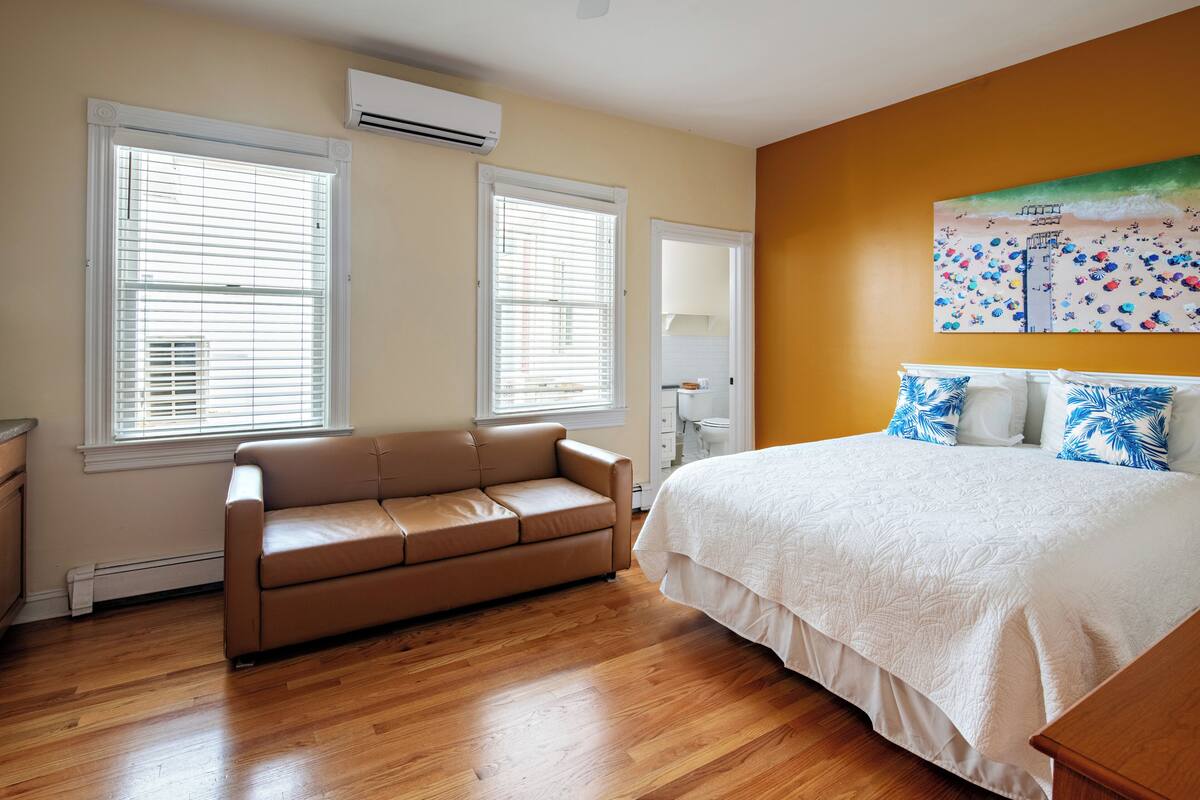 A bedroom with a bed, a sofa, and two windows with blinds.