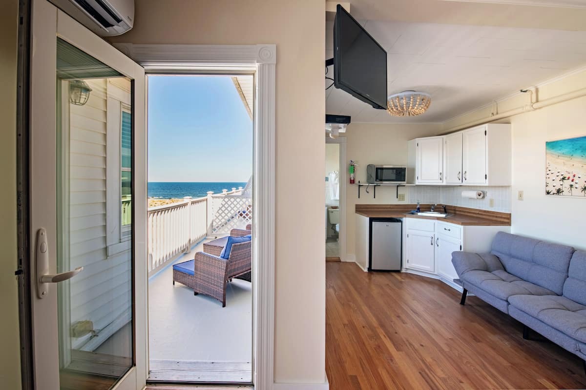 A room with a view of the ocean, a balcony with wicker furniture, a kitchen with white cabinets, and a gray sofa.