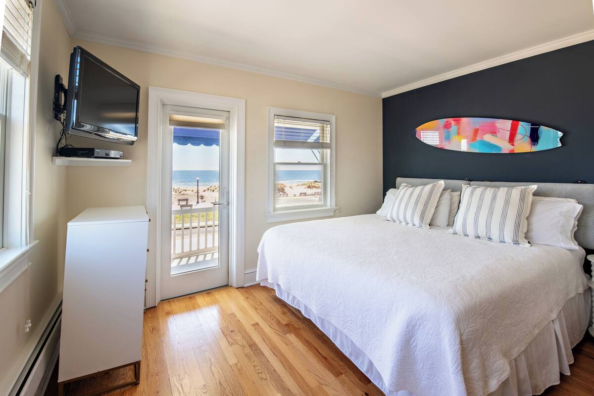 A bedroom with a bed, a TV mounted on a shelf, a window with a view of the beach, and a wall-mounted artwork.