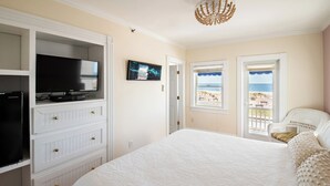A bedroom with a large bed, a TV on a built-in shelf, a painting, and a view of the beach through a window. - Shawmont Hotel  (Ocean Grove)