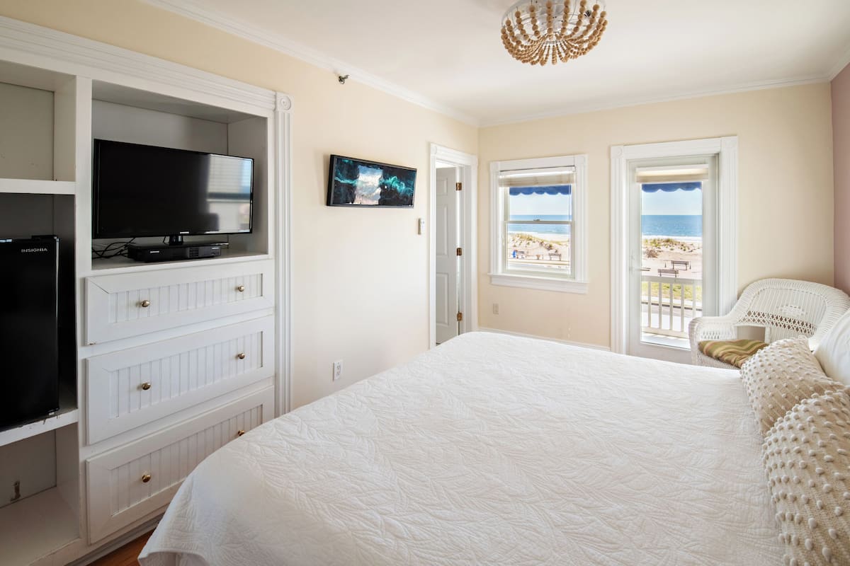 A bedroom with a large bed, a TV on a built-in shelf, a painting, and a view of the beach through a window.