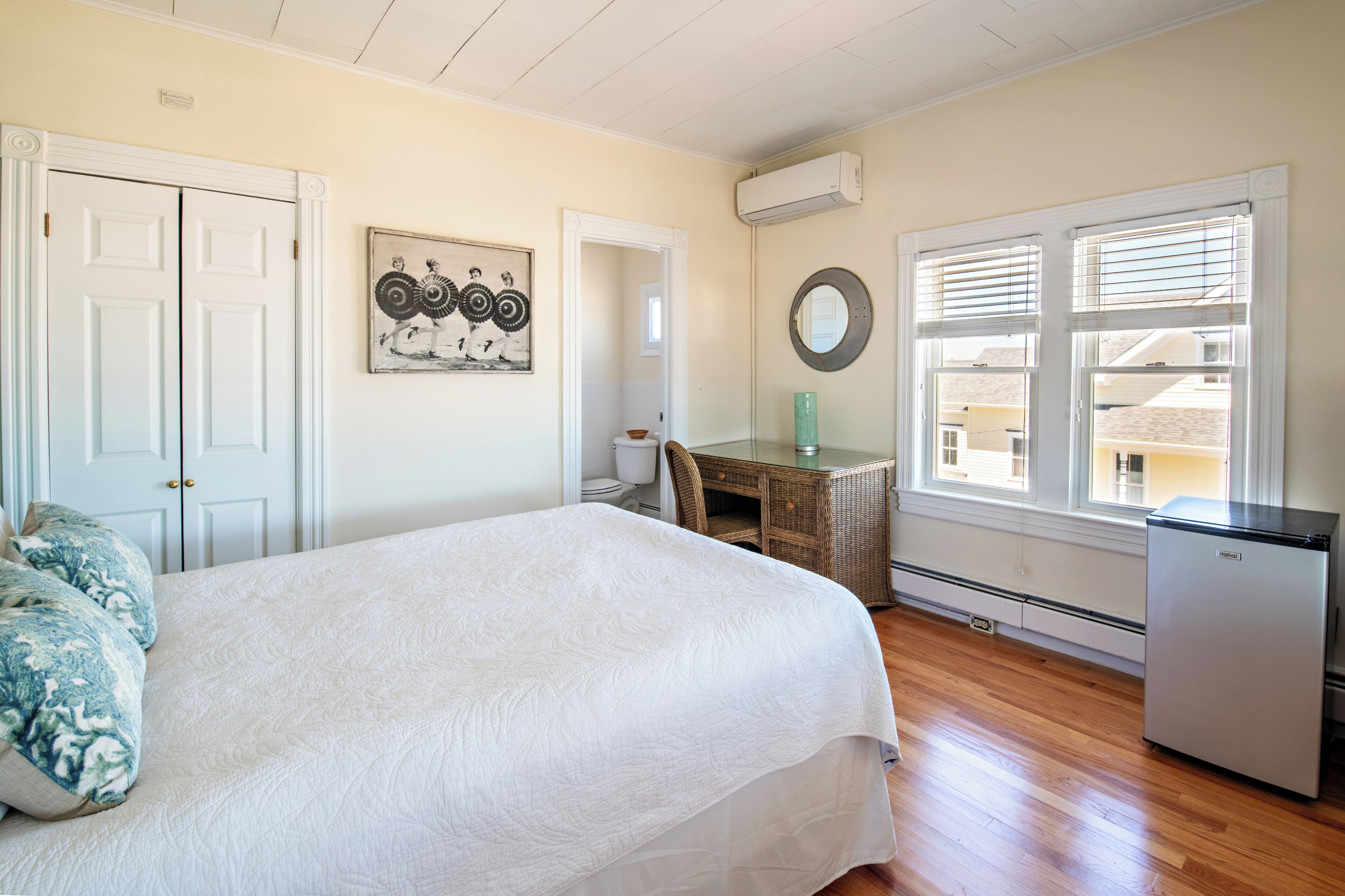 A bedroom with a bed, a desk, a refrigerator, a window with blinds, and a door.